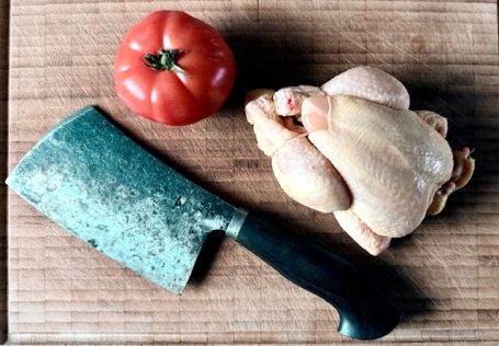 Ein Holztisch mit einem Huhn, einer Tomate und einem Hackmesser.
